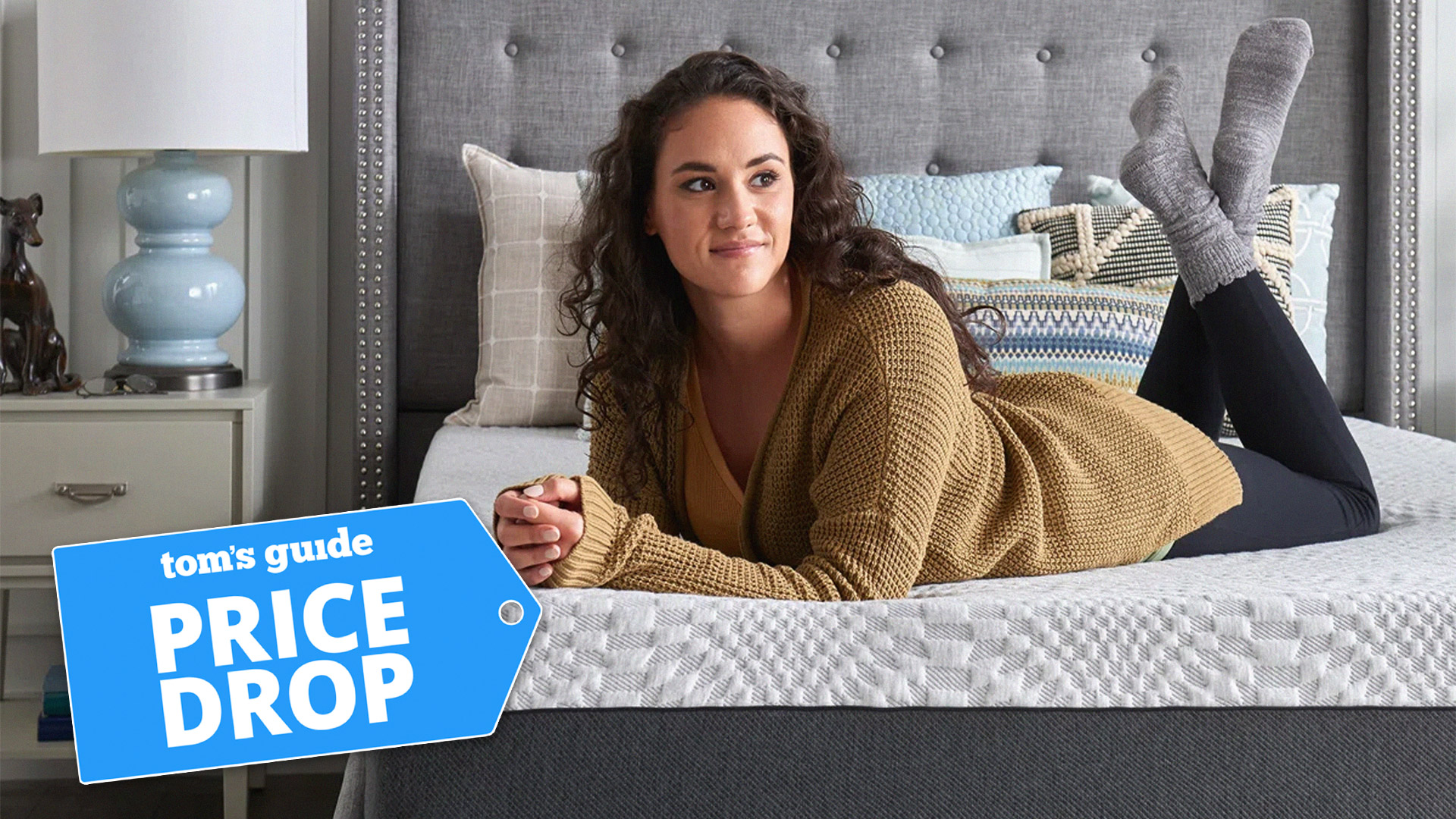 A woman lying on a Sealy Cool 12" Medium Memory Foam Mattress, with a Tom's Guide badge overlaid saying "PRICE DROP"