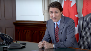 justin trudeau smiling with a canadian flag behind him. he is sitting in a desk chair with a phone in front of him