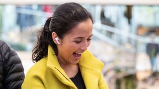 Keltaisessa takissa oleva nainen hymyilee ja kuuntelee Apple AirPods Prolla