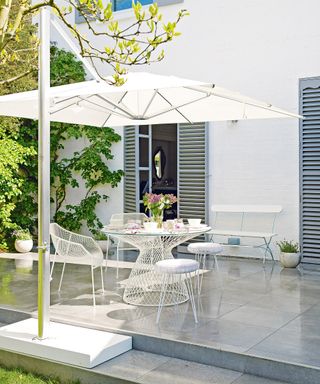 A gray patio which is wet in the rain with a white parasol and white outdoor furniture