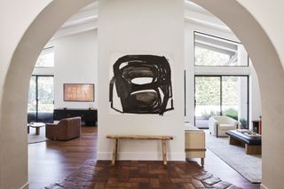 An archway-framed view of an open-plan living space with neutral walls and wooden floor. A black and white abstract artwork hangs on a wall in the centre