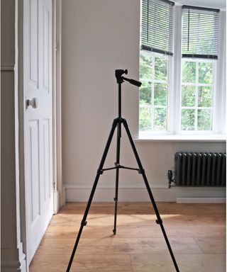 A large tripod in room with white door, wooden flooring, large windows with blinds and radiator