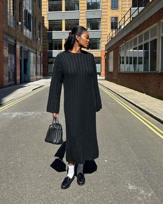 British influencer Marilyn Nwawulor-Kazemaks poses on a street in London wearing a cable-knit maxi sweater dress, Hermès bag, white crew socks, and black loafers