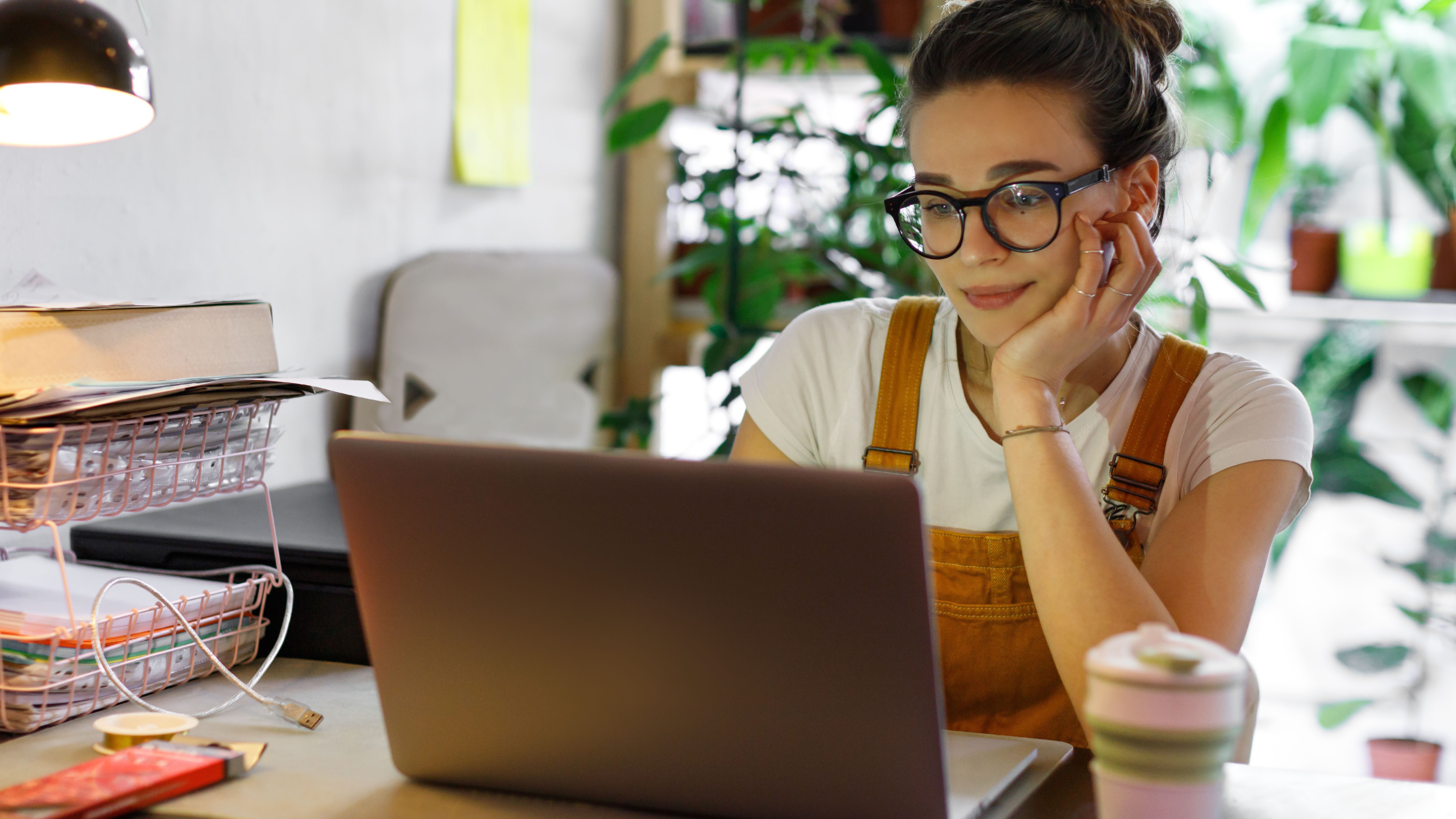 Uma mulher está sentada em uma mesa usando um MacBook e há várias plantas de interior ao fundo