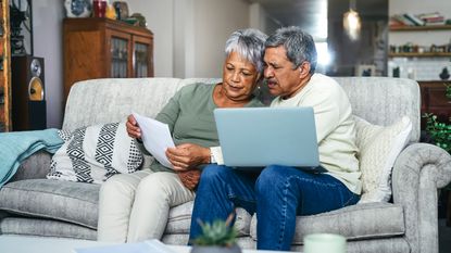 An older couple on a sofa go over paperwork and have a laptop open.
