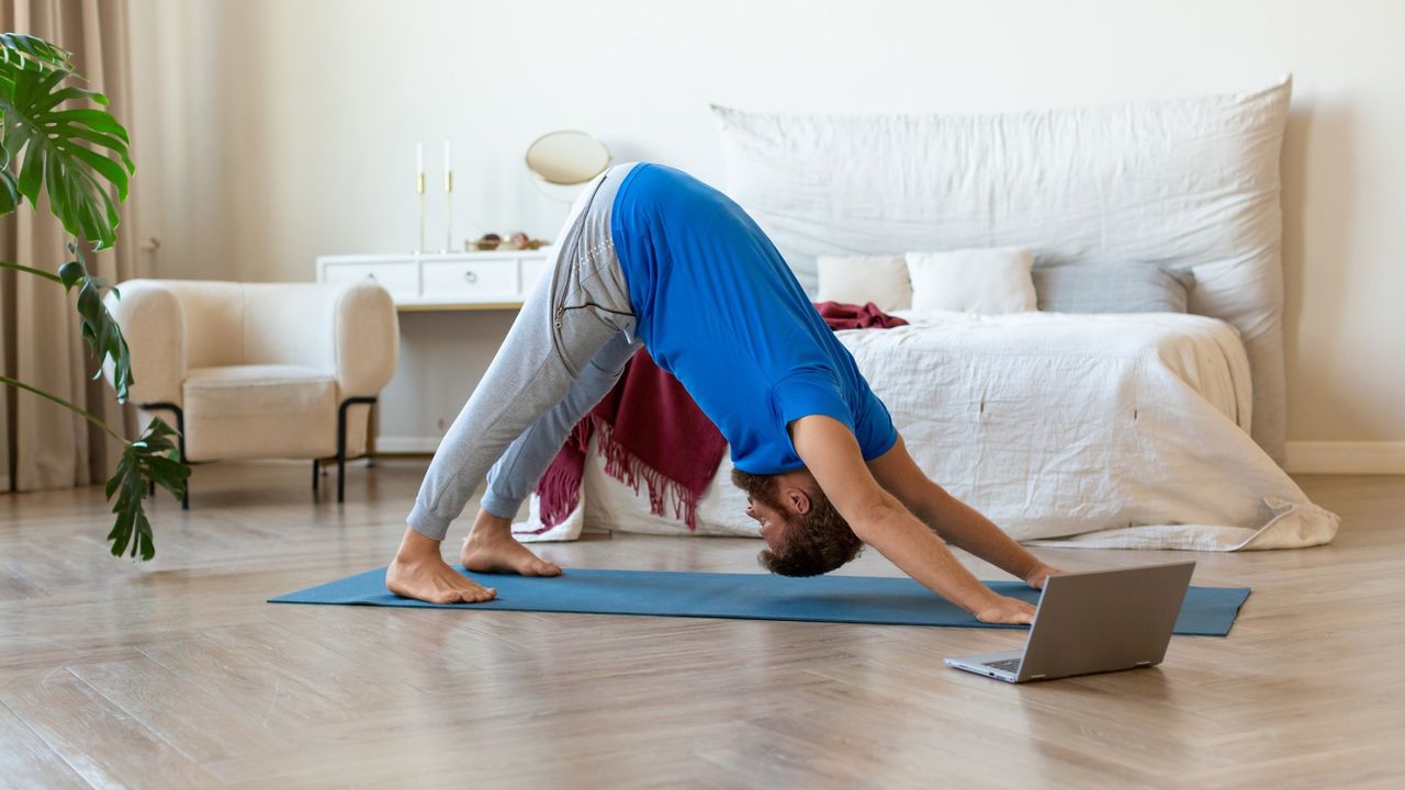 man in a bedroom doing a downward facing dog yoga pose in front of his bed on a yoga mat with a laptop in front of him