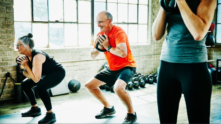 An older woman and man holding a kettlebell in their chest as they squat down in gym class