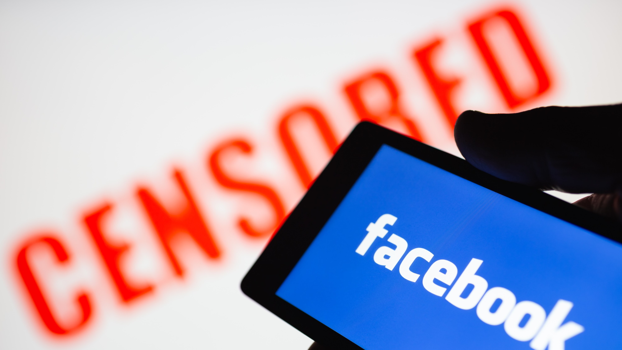 Smartphone en main affichant le logo de Facebook.  Texte censuré rouge flou sur fond
