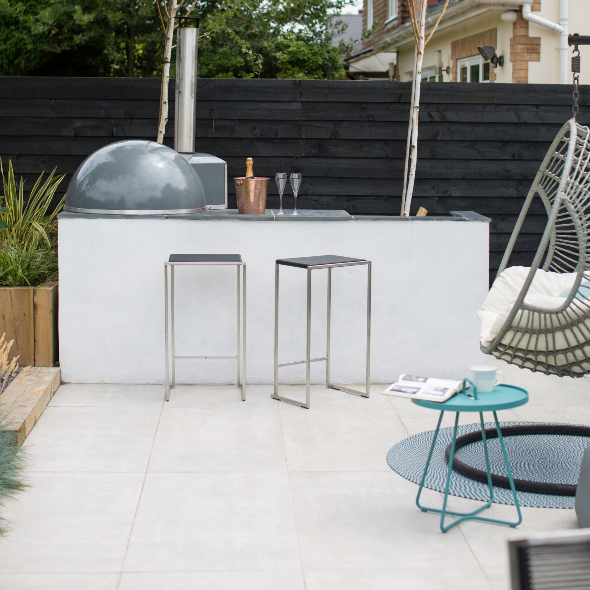 outdoor kitchen with white base, grey worktop, BBQ, white floor tiles, hanging chair, turquoise side table, rug, bar stools