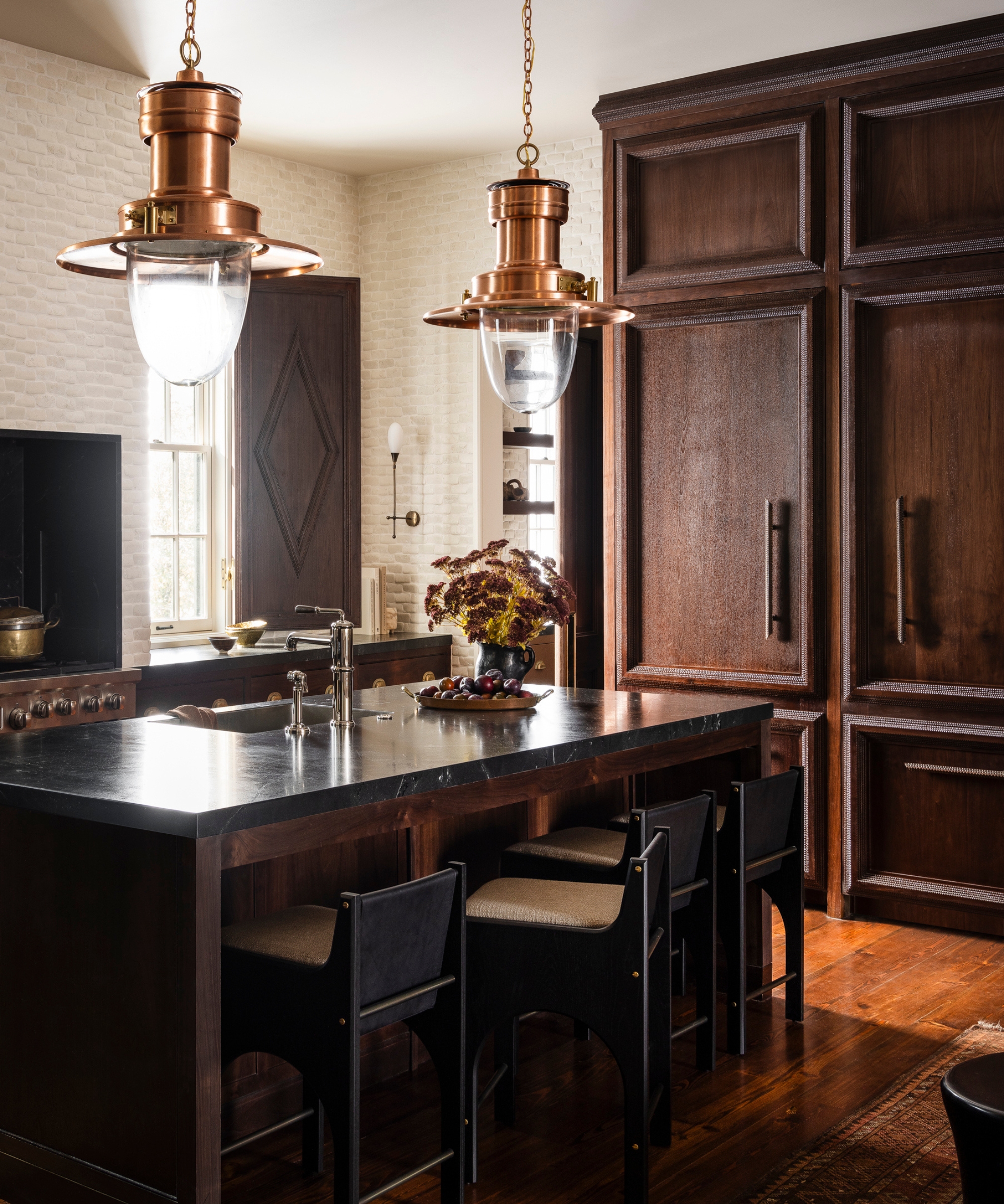 A moody kitchen with dark wood cabinets, black countertops, brass pendant lights, and low-back counter stools