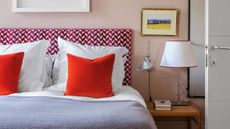 A modern and eclectic bedroom corner features a plush bed with a bold magenta and white chevron headboard and two vibrant orange velvet throw pillows. The soft pink walls are accented by a silver swing-arm reading light and a unique table lamp with a curved wire base sitting on a wooden nightstand. To the right, a white door stands slightly ajar, while a small framed landscape painting and a larger white-framed piece add artistic detail to the cozy, well-lit space.