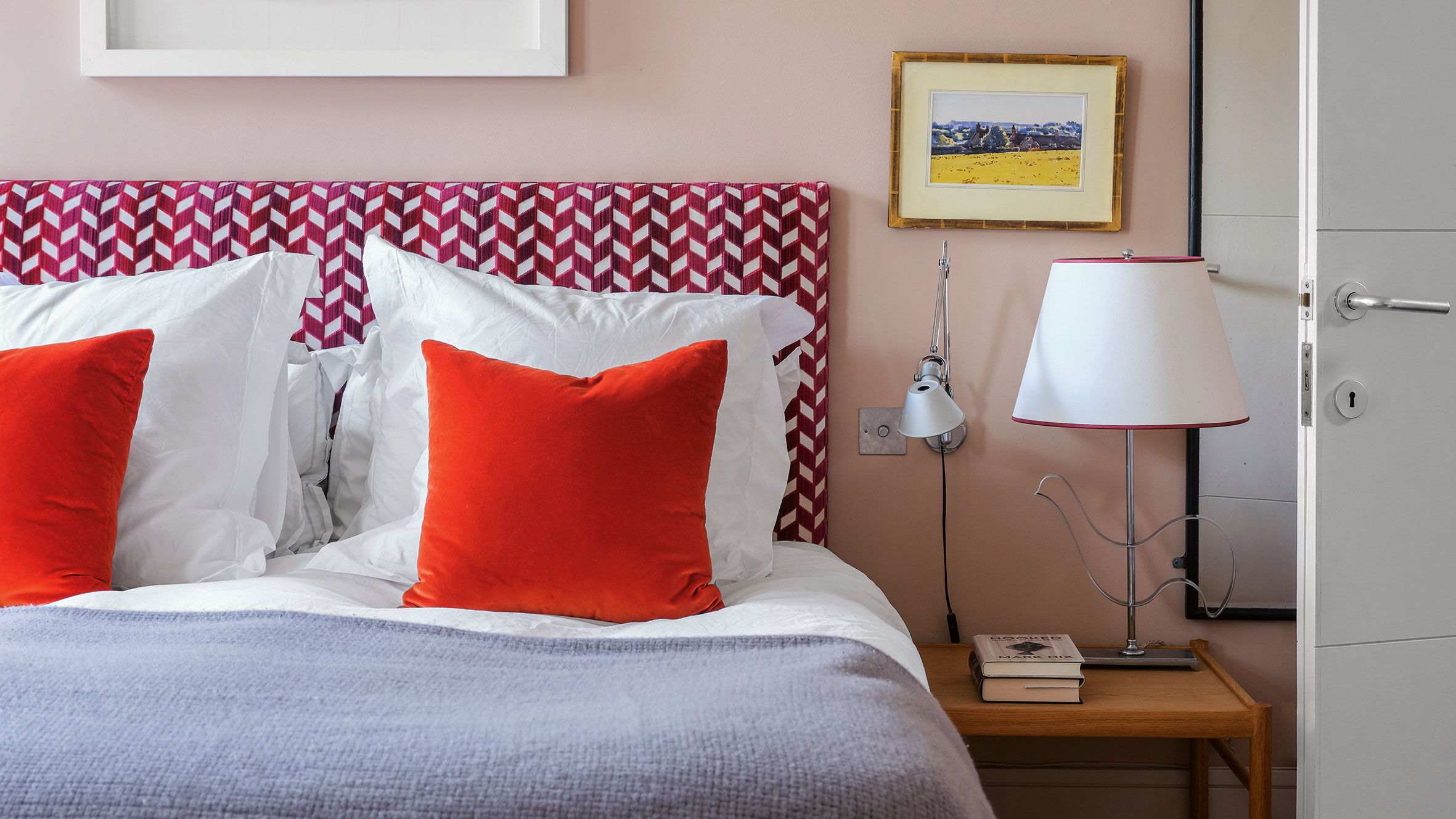 A modern and eclectic bedroom corner features a plush bed with a bold magenta and white chevron headboard and two vibrant orange velvet throw pillows. The soft pink walls are accented by a silver swing-arm reading light and a unique table lamp with a curved wire base sitting on a wooden nightstand. To the right, a white door stands slightly ajar, while a small framed landscape painting and a larger white-framed piece add artistic detail to the cozy, well-lit space.