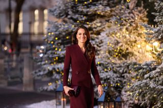 Kate Middleton walking past snow-covered lit up Christmas trees wearing a long coat and smiling