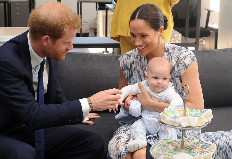 Prince Harry has red hair and wears a suit and holds the hand of his new baby, Archie, who sits on wife Meghan Markle's lap