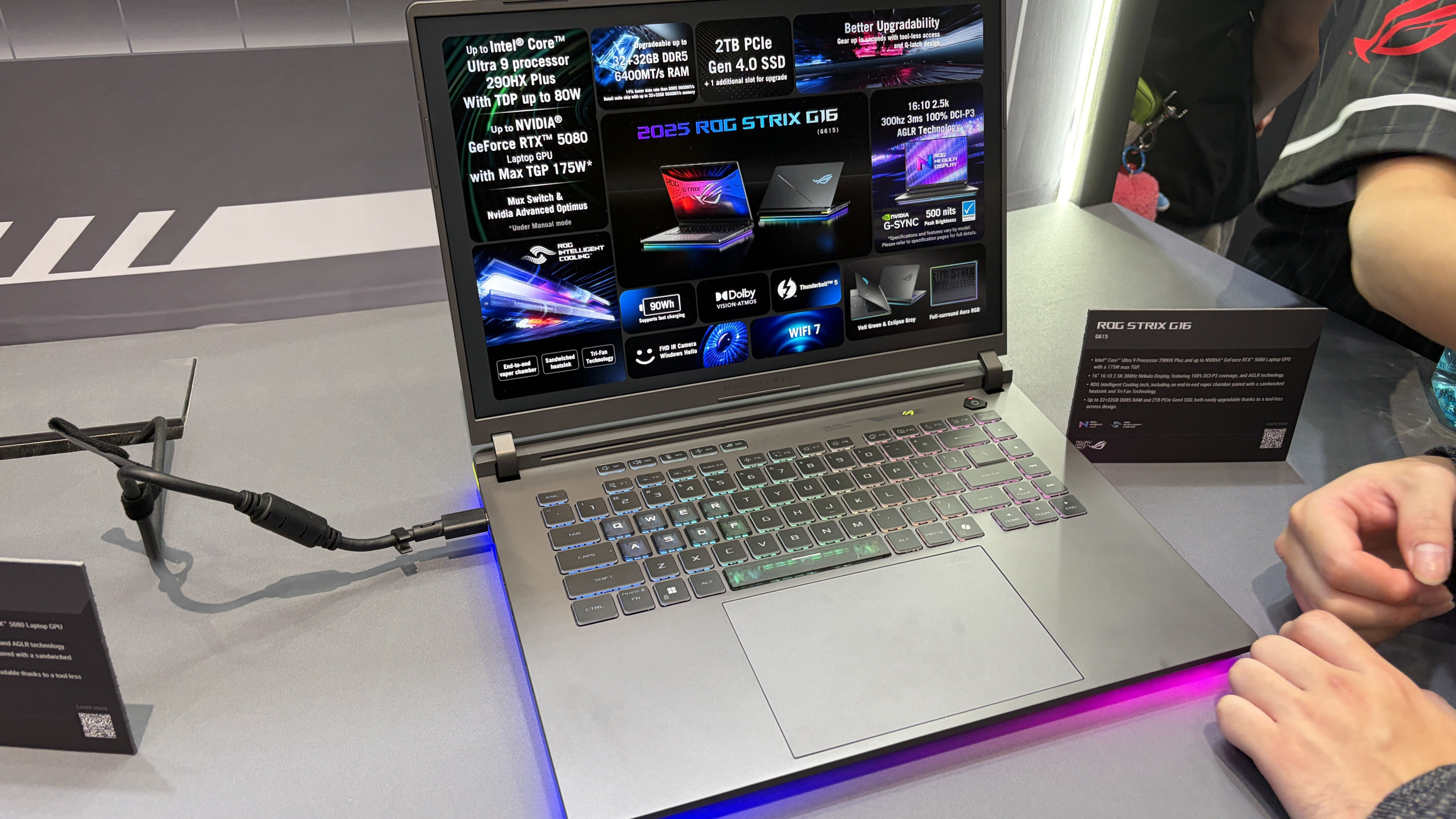 An ASUS ROG Strix G16 laptop with an RGB backlit keyboard displaying promotional specifications on its screen, connected to a power cable with information cards next to it.