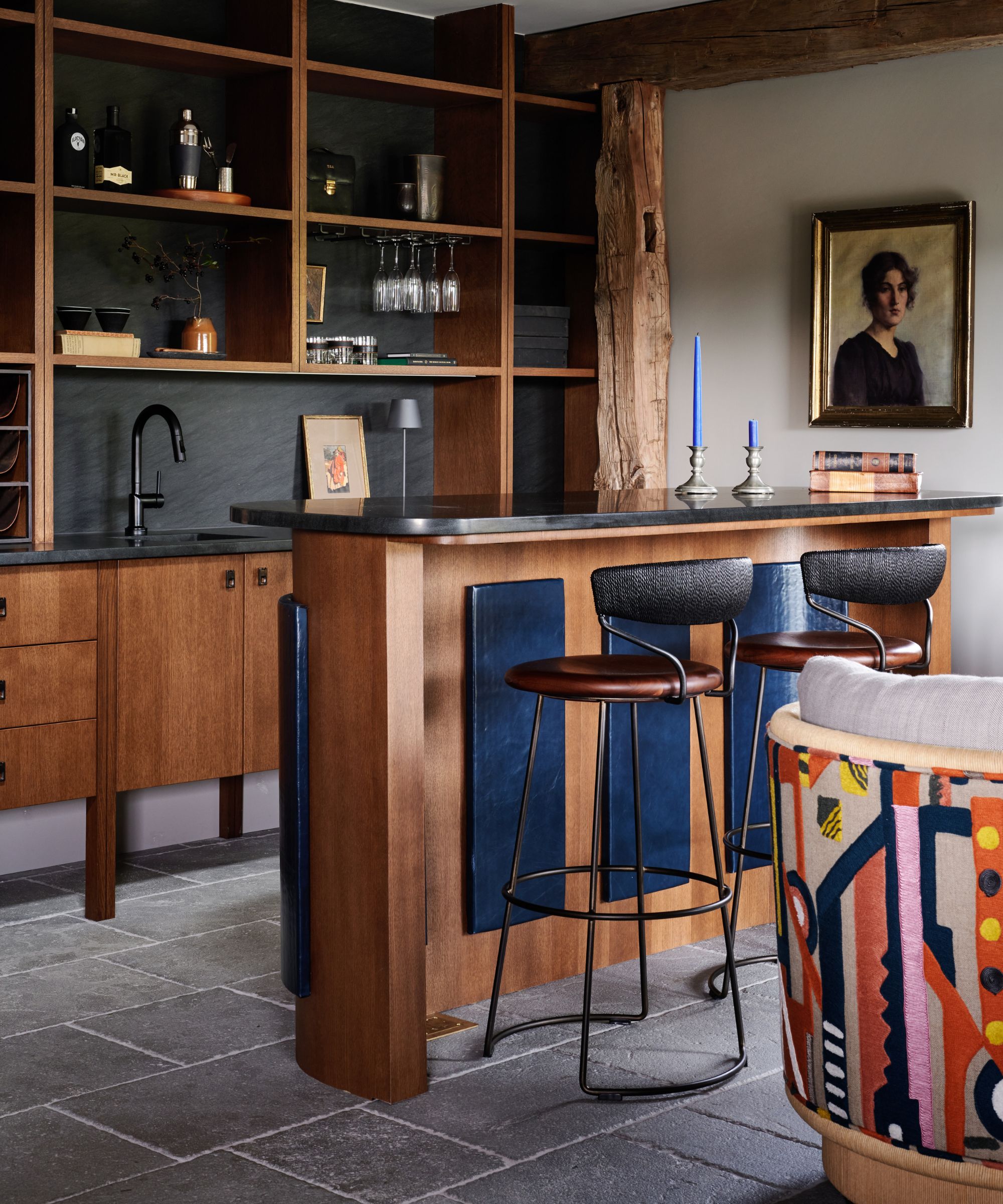 Wooden kithcen with slim wooden bar island, bar stool seating, stone floor, artwork on wall in background, patterned chair in foreground