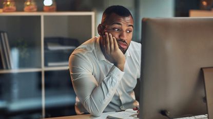 A worried-looking man, chin in hand, sits at his desk and looks at his computer monitor.