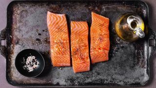 oily salmon on a baking tray next to a bottle of olive oil and a bowl of seasoning