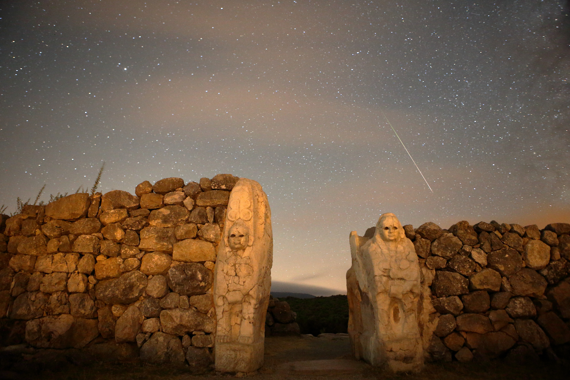 Una scia di meteoriti prominente nel cielo sopra un muro di pietra con due figure scolpite nella roccia.
