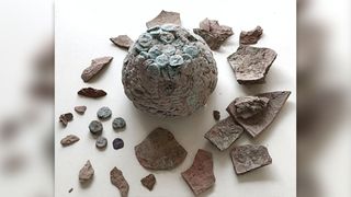 Hoard of copper coins tarnished green and fused together into a single lump with five additional coins and the cracked remains of its container on a white display.