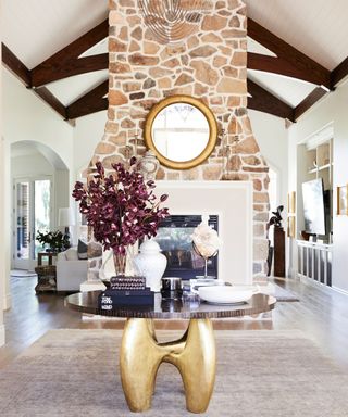 Large living room with double height ceilings and vaulted ceiling with dark wooden beams, feature stone fireplace, light wooden flooring, large gray rug, round glass table with gold legs, decorated with vases, ornaments and books