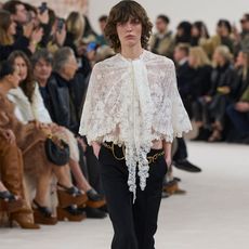 Chloe model wearing white lace blouse, gold belt, and black trousers walking on the runway