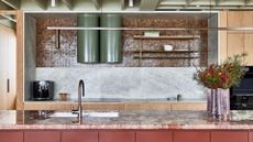 A stone kitchen with a tiled overhead accent wall, green tubular exhaust pipes, a pink marbled kitchen countertop, brown brassy hardware, a vase of flowers, and a coffee machine