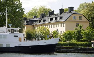 Hotel Skeppsholmen, Stockholm. A large three storey beige building with a black roof in front of a river with a large boat on it.