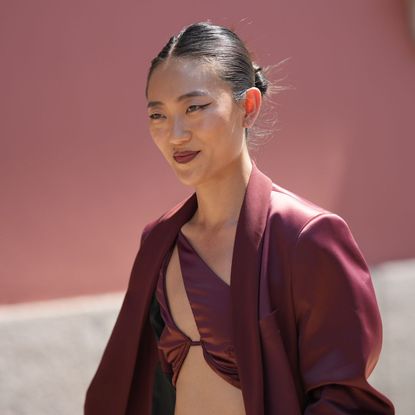 best brown eyeliners - woman smiling wearing a burgundy top and blazer with a chignon and a sharp brown eyeliner flick and burgundy lip - gettyimages 1499115748