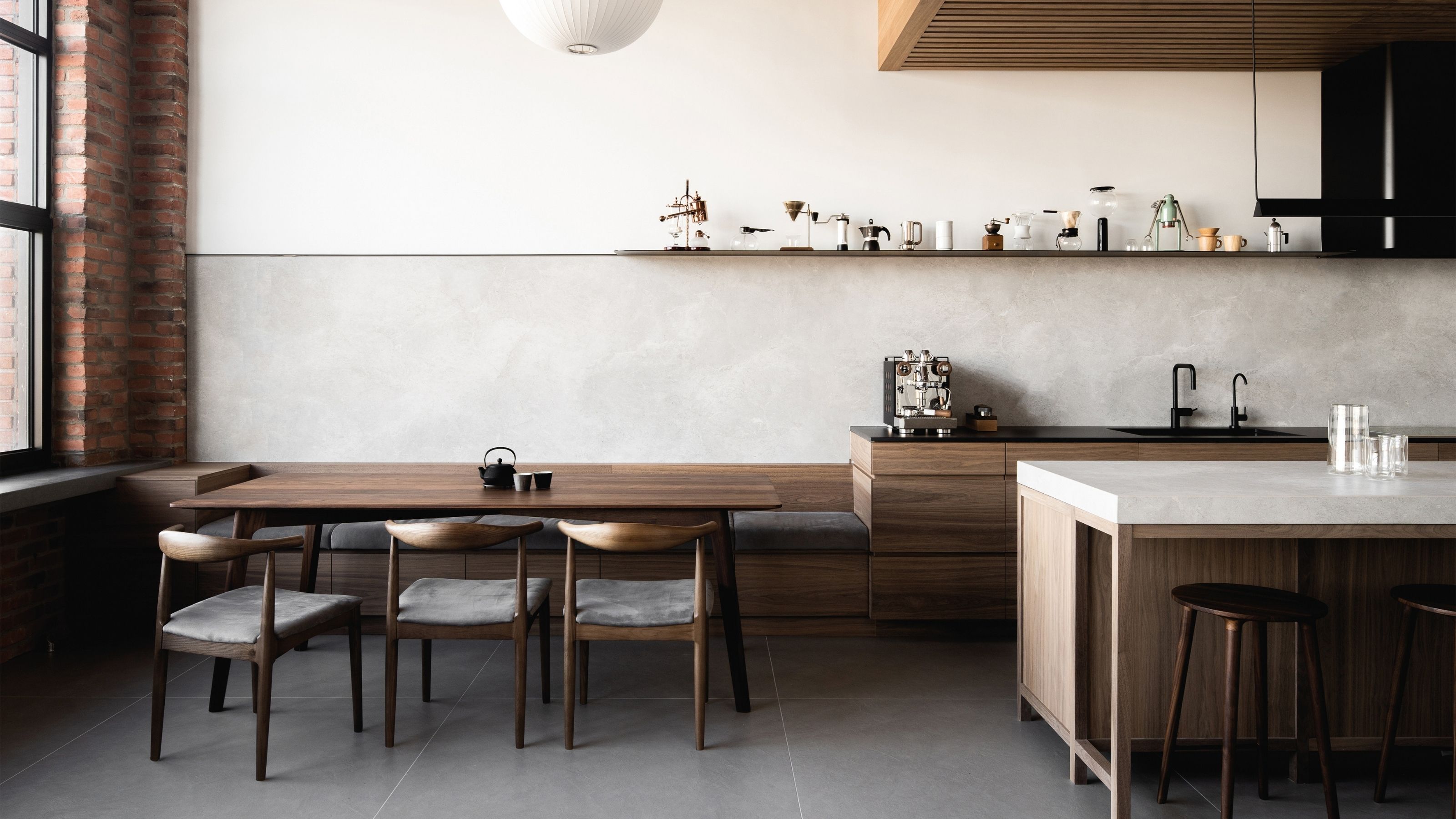 Image of a neutral, open kitchen with lots of sunlight. There is love wooden cabinetry, a wooden island with a marble top, and lots of different coffee makers in the space.