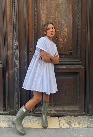 a photo of a summer rainy day outfit with a woman wearing a dainty earrings, a white minidress, and green rain boots