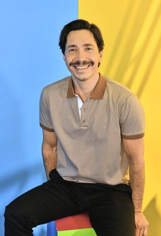 justin long sits on a stool wearing a brown shirt and black pants smiling in a promo shot for life is short