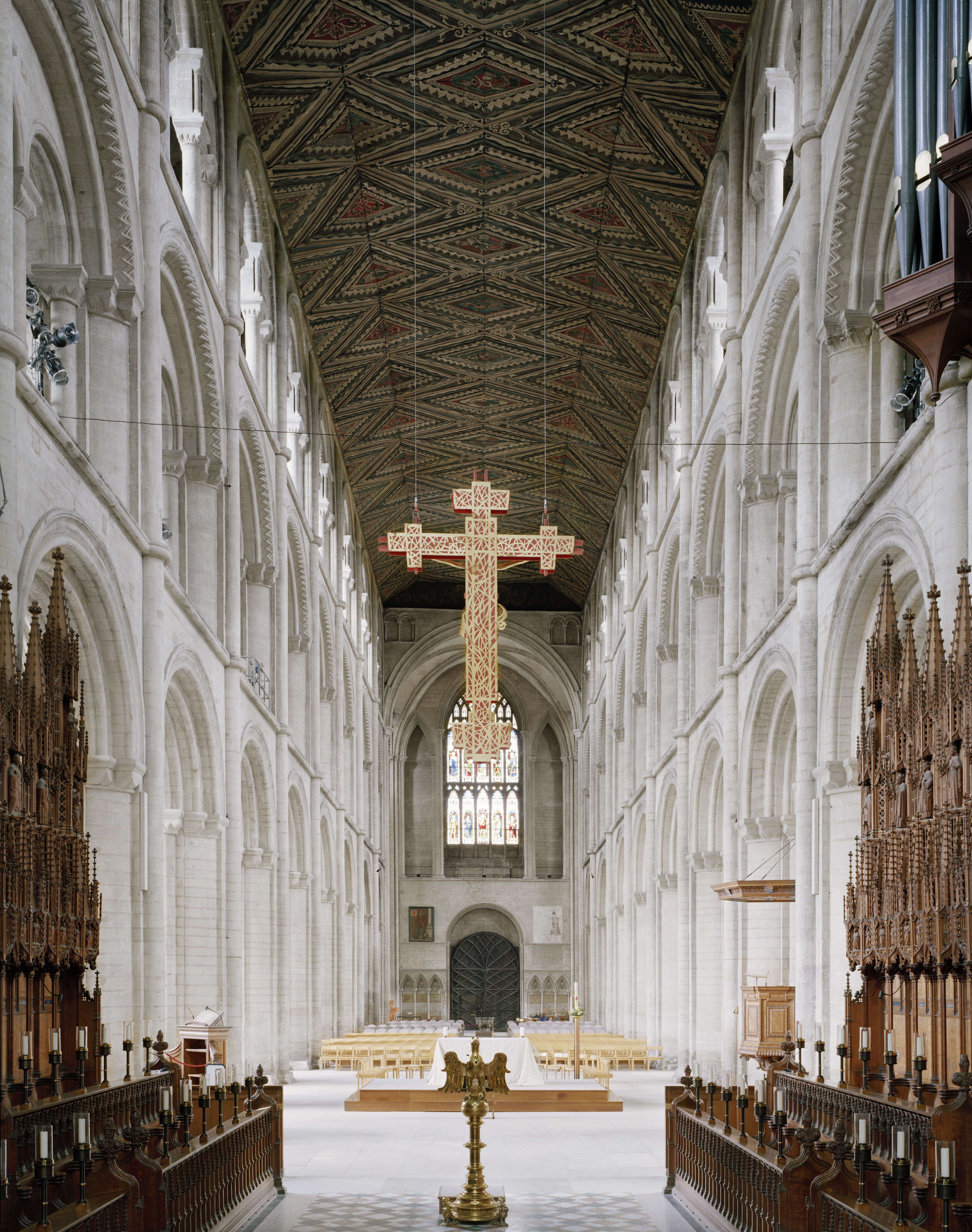 The nave of Peterborough Cathedral, with a large wooden cross suspended beneath its painted diamond-patterned ceiling above the choir stalls.