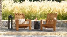 Two Teak Outdoor Adirondack Lounge Chairs with a small black side table in between with drinks and a large black lantern next to the chair on the left with a wall of ornamental grasses behind