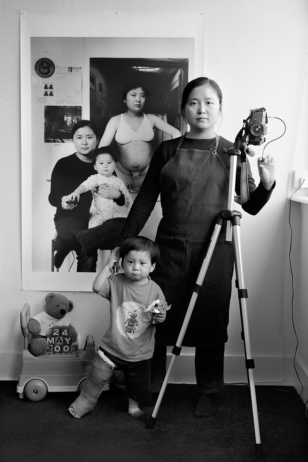 A black-and-white photograph showing a mother and her young child with a leg cast standing next to a camera on a tripod, positioned in front of layered studio portraits tracking the child's growth.