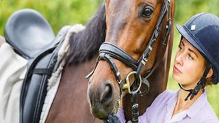 A horse rider standing with their face close to a brown horse's face wearing a saddle and reigns