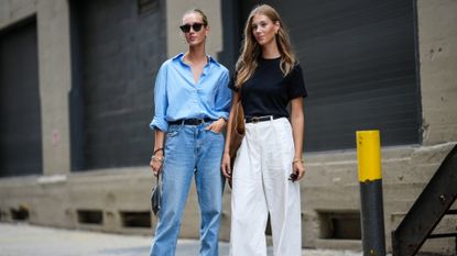 A woman wears a blue button-down shirt and blue jeans and another women wears a black tee shirt and white pants.