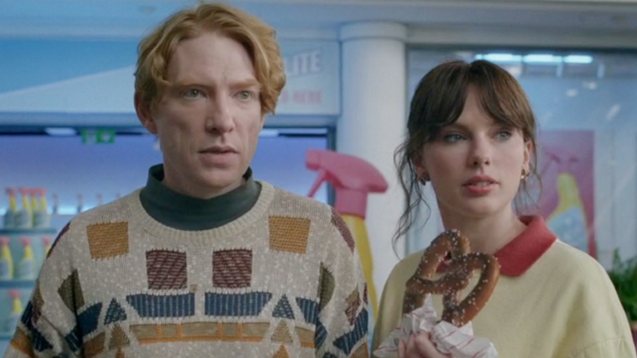 From left to right: Domhnall Gleeson and Taylor Swift holding a pretzel in the Opalite music video. 