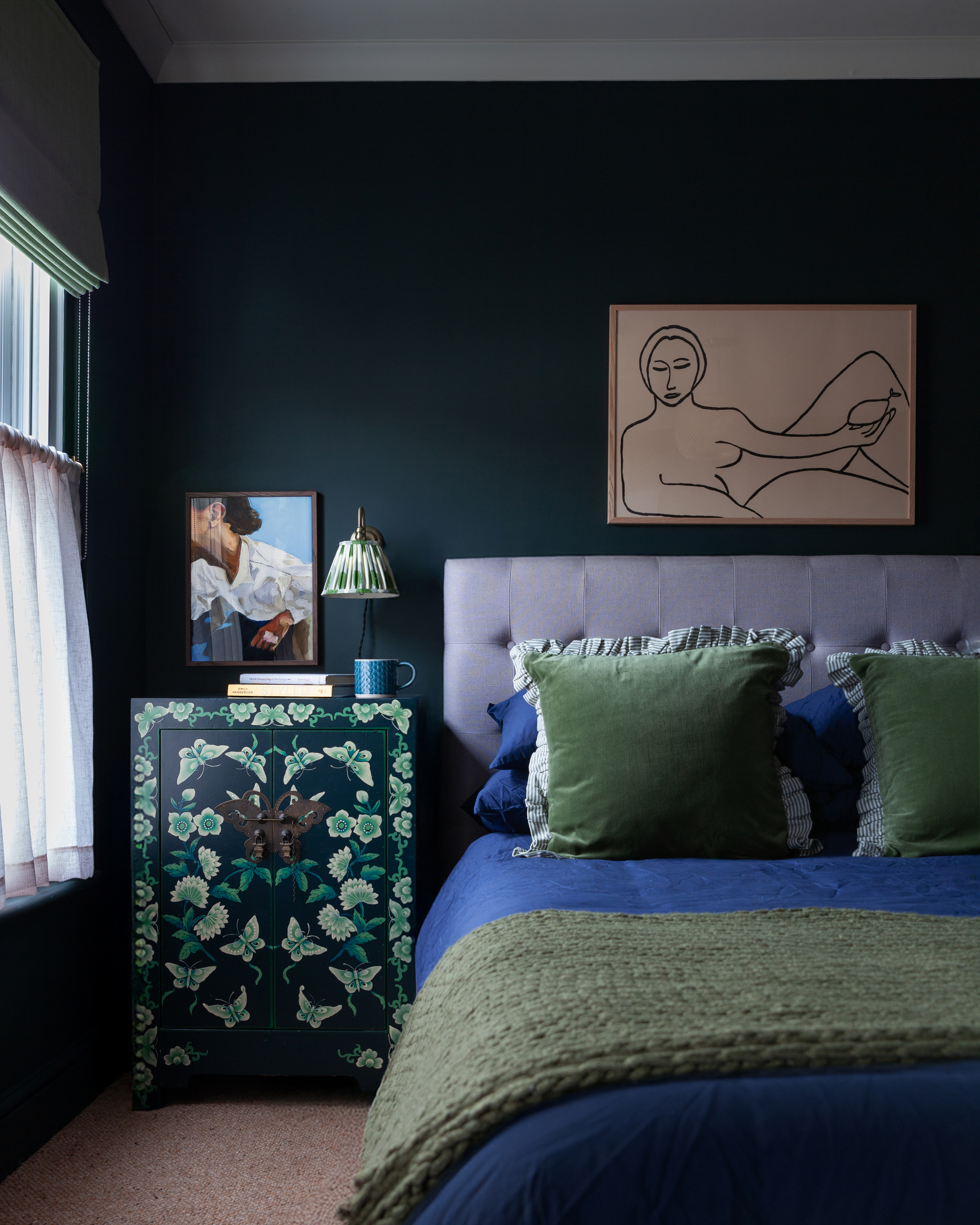 A green bedroom with blue accents, blue bedding, and hand-painted bedside table, green cushions, sheer curtains, and an artwork hanging over the bed
