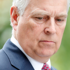 A closeup picture of Prince Andrew looking upset and wearing a blue suit with a white dress shirt and red tie