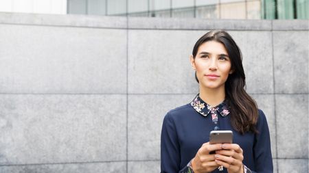 A young woman holding a smartphone looks up thoughtfully.