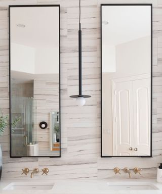 Two tall rectangular bathroom mirrors with black borders on a gray marble wall and a long black pendant light between them, with two sets of gold faucets and white sinks below them