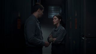 A man and woman (both wearing grayish-blue jumpsuits) are having a conversation in a small corridor. The woman has her arms crossed.