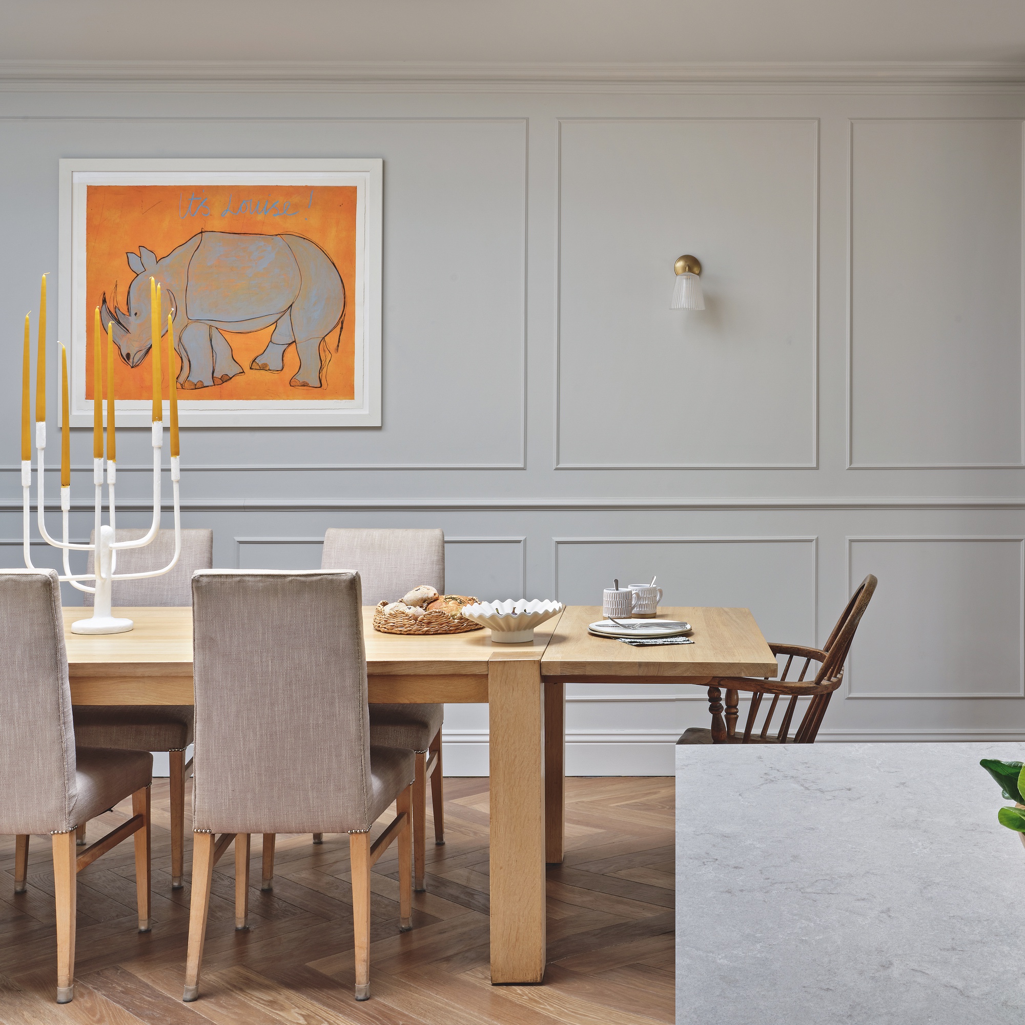 dining area in open-plan kitchen with panel walls painted grey, large wooden dining table, grey upholstered dining chairs and colourful artwork of a rhino on an orange background