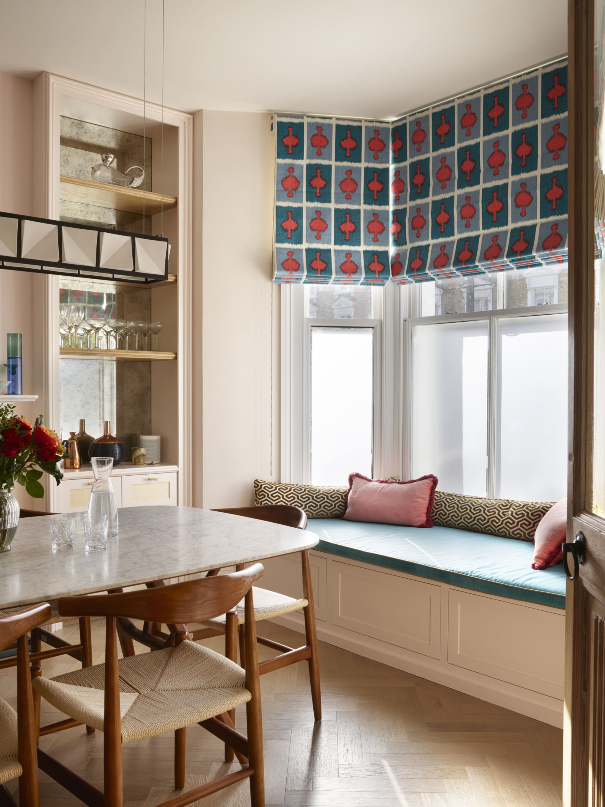 Image of a bay window with patterned roman shades and a teal bench seat in a light pink dining room with a marble dining table that has wood and rattan dining chairs around it.