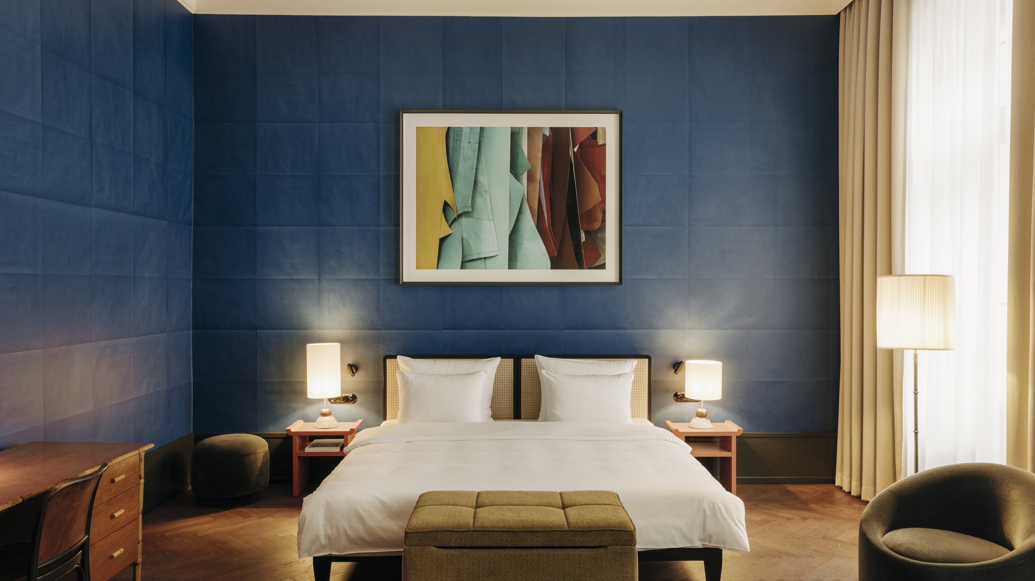 A hotel bedroom with paper-like blue walls with a folded effect, warm bed table lamps and a standing one, green fabric furnishings, wooden pieces, and wooden floors.