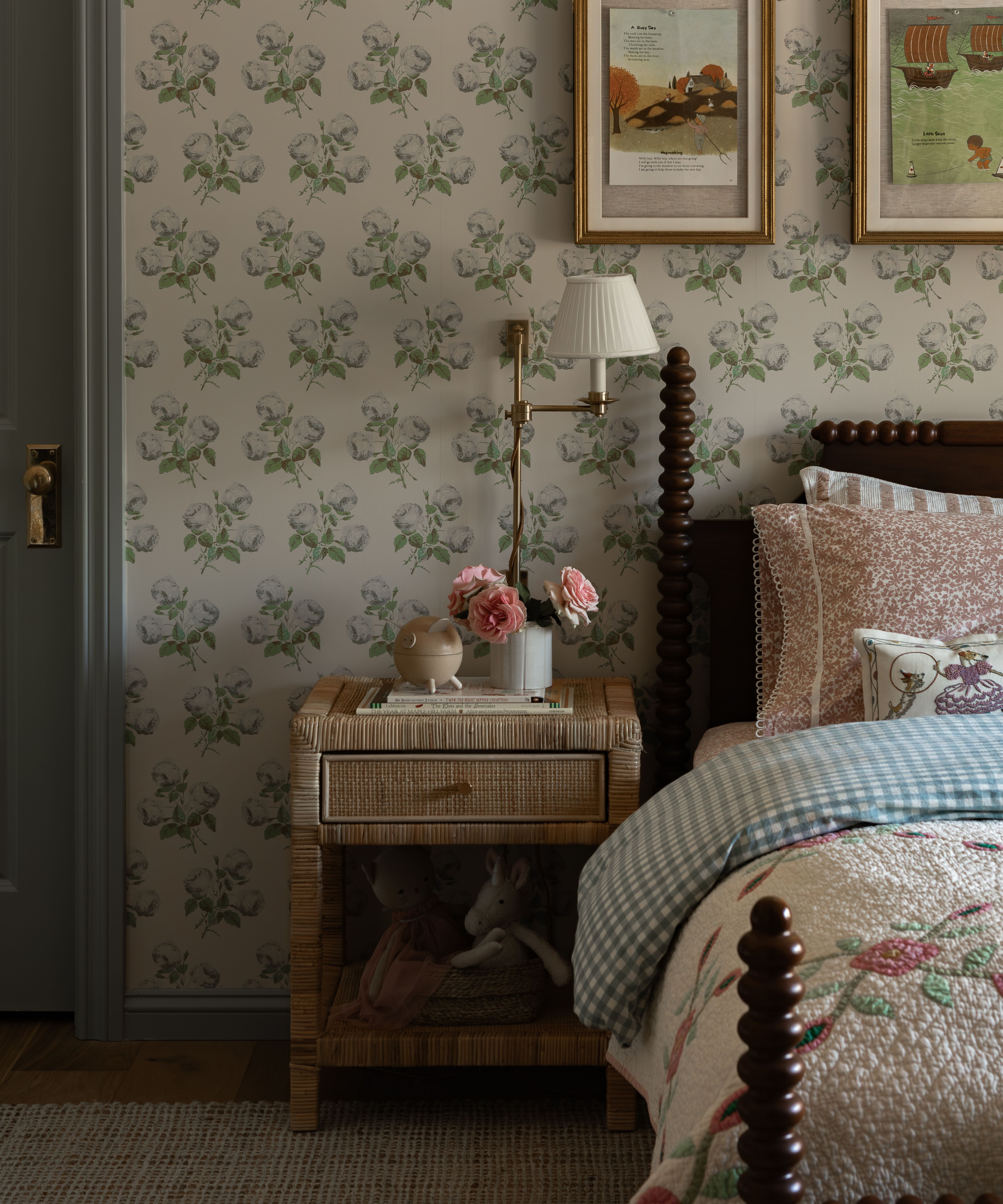 a floral wallpapered child's bedroom with sage green woodwork, a dark wooden bobbin bed with a wicker nightstand and layered gingham and floral bedding