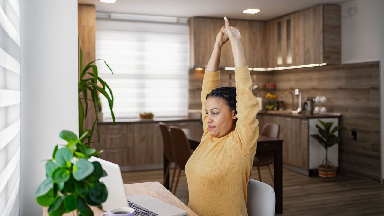 Woman sitting down at desk with laptop, reaching arms above her head to stretch, using one of the best stretching apps