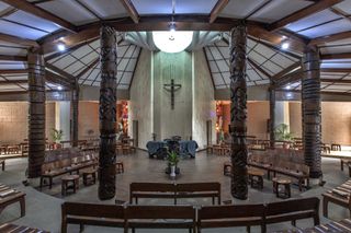 Interior image of the Dominican chapel in Ibadan and its 12 carved columns that reference the 12 disciples, stone floor, wooden beam ceiling, wooden pillars, wooden benches, white vase and plant in the centre, white centre piece wall with mounted religious cross, brown walls, table covered with blue cloth, chairs, potted plants