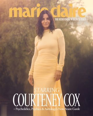 Courteney Cox standing against a backdrop of trees in a full-length yellow dress and smiling